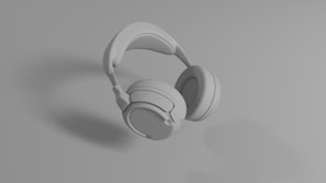 A set of over-ear headphones in a monochromatic gray color scheme, resting on a smooth surface. The design appears sleek and modern with cushioned ear pads and an adjustable headband.