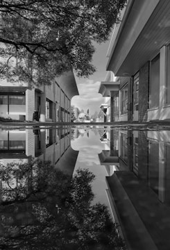 Black and white street scene capturing a lone figure reflected in a rain puddle on a quiet Indian alley.