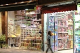 A vibrant and colorful storefront features an assortment of dried fruits, nuts, and snacks. The display shows neatly arranged goods in jars and containers. Adjacent to this is a toy store section filled with boxed dolls and various toys, illuminated by bright lights. A person is walking past the store, carrying a shopping bag.