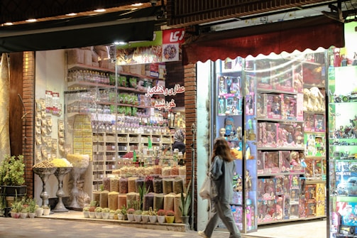 A vibrant and colorful storefront features an assortment of dried fruits, nuts, and snacks. The display shows neatly arranged goods in jars and containers. Adjacent to this is a toy store section filled with boxed dolls and various toys, illuminated by bright lights. A person is walking past the store, carrying a shopping bag.