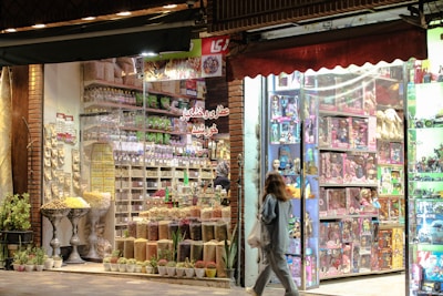 A vibrant and colorful storefront features an assortment of dried fruits, nuts, and snacks. The display shows neatly arranged goods in jars and containers. Adjacent to this is a toy store section filled with boxed dolls and various toys, illuminated by bright lights. A person is walking past the store, carrying a shopping bag.
