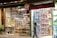 A vibrant and colorful storefront features an assortment of dried fruits, nuts, and snacks. The display shows neatly arranged goods in jars and containers. Adjacent to this is a toy store section filled with boxed dolls and various toys, illuminated by bright lights. A person is walking past the store, carrying a shopping bag.