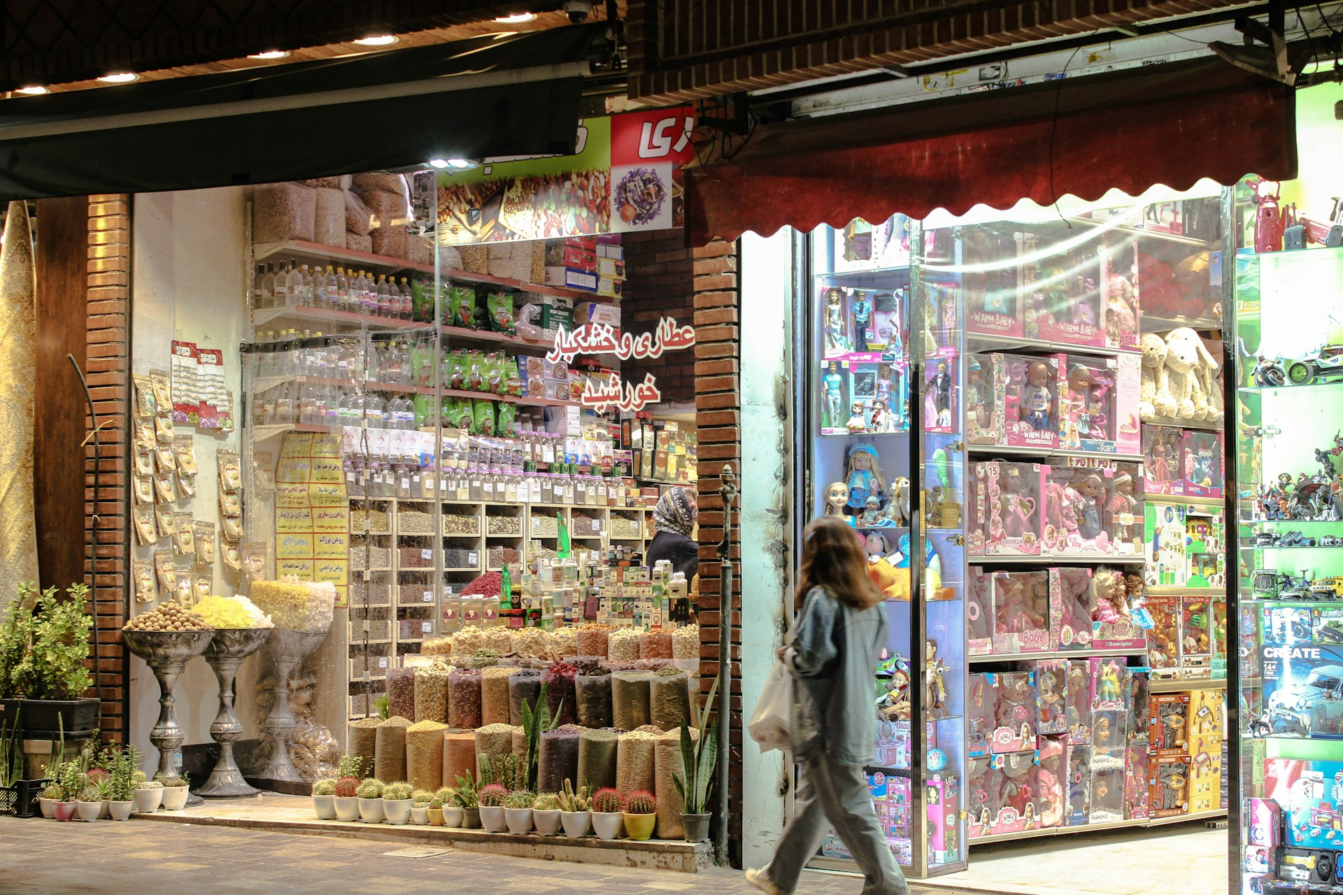 A vibrant and colorful storefront features an assortment of dried fruits, nuts, and snacks. The display shows neatly arranged goods in jars and containers. Adjacent to this is a toy store section filled with boxed dolls and various toys, illuminated by bright lights. A person is walking past the store, carrying a shopping bag.