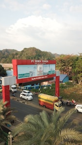 A busy urban scene featuring a large red and blue billboard with vehicles passing underneath. The sign reads PT. Semen Tonasa, set against a background of green hills and partly cloudy skies. Palm trees are in the foreground, adding to the tropical atmosphere.