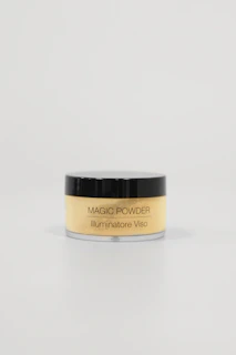 A small, cylindrical container with a black lid and transparent body, labeled 'MAGIC POWDER Illuminatore Viso'. It is filled with a light yellow powder. The container is positioned against a plain, light-colored background, emphasizing minimalistic and clean aesthetics.