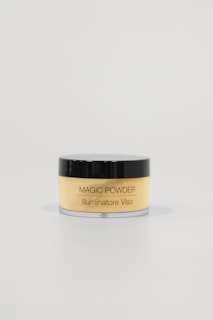 A small, cylindrical container with a black lid and transparent body, labeled 'MAGIC POWDER Illuminatore Viso'. It is filled with a light yellow powder. The container is positioned against a plain, light-colored background, emphasizing minimalistic and clean aesthetics.