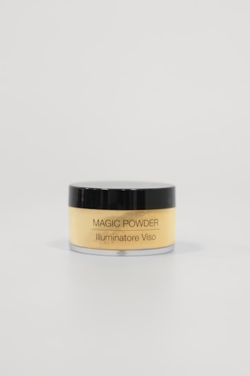 A small, cylindrical container with a black lid and transparent body, labeled 'MAGIC POWDER Illuminatore Viso'. It is filled with a light yellow powder. The container is positioned against a plain, light-colored background, emphasizing minimalistic and clean aesthetics.