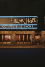 Neon lights illuminate the sign of a music hall with the words 'Music Hall' in elegant cursive. Beneath it, another sign announces upcoming shows on October 7th and 8th. The building's entrance features large glass doors and a reflective surface, hinting at an evening or nighttime setting.