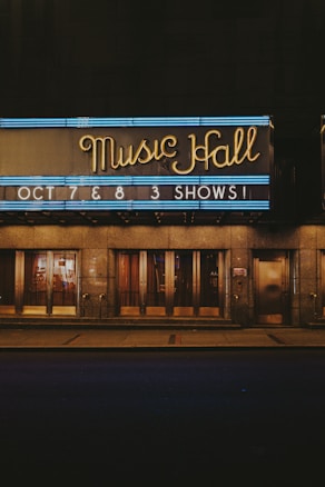 Neon lights illuminate the sign of a music hall with the words 'Music Hall' in elegant cursive. Beneath it, another sign announces upcoming shows on October 7th and 8th. The building's entrance features large glass doors and a reflective surface, hinting at an evening or nighttime setting.