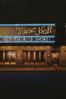 Neon lights illuminate the sign of a music hall with the words 'Music Hall' in elegant cursive. Beneath it, another sign announces upcoming shows on October 7th and 8th. The building's entrance features large glass doors and a reflective surface, hinting at an evening or nighttime setting.