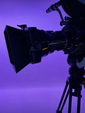 A close-up view of a professional film camera, focusing on the lens and matte box, set against a vibrant purple background. The camera is mounted on a tripod, and the lighting emphasizes the camera's intricate details and mechanisms.