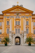 An ornate baroque-style building with a symmetrical facade features a prominent clock and decorative sculptures under a triangular pediment. The walls are painted in warm yellow and white hues. A grand archway at the center leads to an inner courtyard, and two potted palm trees flank the entrance. The architecture is detailed with columns and large windows.