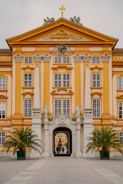 An ornate baroque-style building with a symmetrical facade features a prominent clock and decorative sculptures under a triangular pediment. The walls are painted in warm yellow and white hues. A grand archway at the center leads to an inner courtyard, and two potted palm trees flank the entrance. The architecture is detailed with columns and large windows.