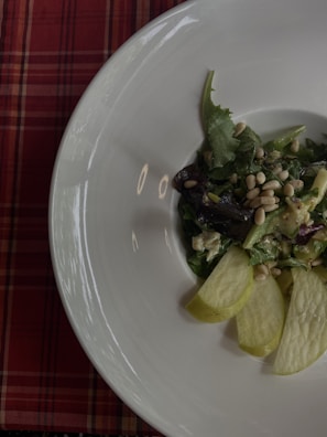 A plate with a fresh salad containing leafy greens, pine nuts, and slices of green apple, placed on a white dish. The background features a red plaid tablecloth.