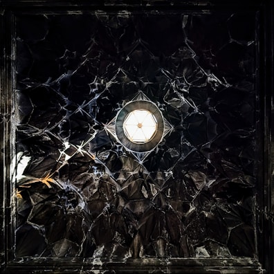 A geometric, textured ceiling with a central hexagonal light fixture. The dark surface has an intricate, reflective pattern that resembles a mosaic of mirrored tiles.