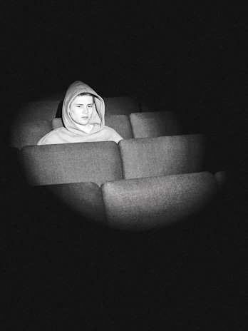 A person wearing a hoodie is seated alone in an empty row of theater-style seats. The scene is in black and white, with shadows adding a dramatic effect. The person appears to be in deep thought or contemplation.