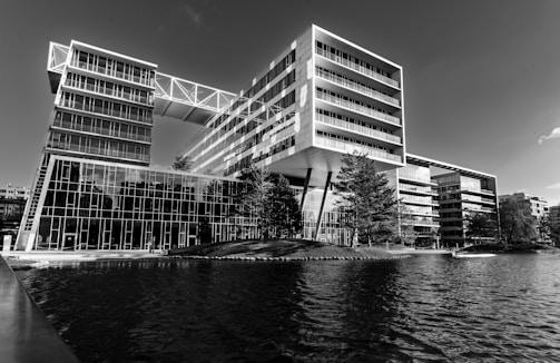 A modern architectural building with large glass windows situated alongside a body of water. The building has a dynamic design with various angular and rectangular elements, including overhanging sections supported by pillars. The landscape around it features several trees and a small grassy area.