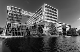 A modern architectural building with large glass windows situated alongside a body of water. The building has a dynamic design with various angular and rectangular elements, including overhanging sections supported by pillars. The landscape around it features several trees and a small grassy area.