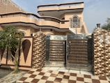 A modern two-story house with a distinct architectural design featuring curved balconies and a combination of beige and brown color tones. The front yard includes a checkered brown and white tiled pathway leading to a large metal gate with wood accents. The exterior walls are adorned with decorative elements and there is a tree and some greenery on the left side.