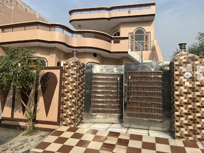 A modern two-story house with a distinct architectural design featuring curved balconies and a combination of beige and brown color tones. The front yard includes a checkered brown and white tiled pathway leading to a large metal gate with wood accents. The exterior walls are adorned with decorative elements and there is a tree and some greenery on the left side.