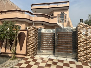 A modern two-story house with a distinct architectural design featuring curved balconies and a combination of beige and brown color tones. The front yard includes a checkered brown and white tiled pathway leading to a large metal gate with wood accents. The exterior walls are adorned with decorative elements and there is a tree and some greenery on the left side.