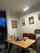A cozy workspace, featuring a wooden desk with various decorative items such as picture frames, a vase, and candles. Above the desk, there are framed artworks on the wall. The space is softly illuminated by a stylish curved floor lamp. The atmosphere feels inviting and personal.