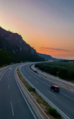 A sunset view of a quiet highway stretching through rolling hills, with a compact car in the distance.