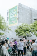 A lively outdoor gathering with many people, including families and children, enjoying themselves in a public space. Soap bubbles float in the air, adding a whimsical touch to the scene. A large colorful banner on a building in the background with the words 'HOW FUN'. People are interacting, some are taking photos, and a child is sitting on an adult's shoulders. The atmosphere is festive and cheerful.
