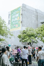 A lively outdoor gathering with many people, including families and children, enjoying themselves in a public space. Soap bubbles float in the air, adding a whimsical touch to the scene. A large colorful banner on a building in the background with the words 'HOW FUN'. People are interacting, some are taking photos, and a child is sitting on an adult's shoulders. The atmosphere is festive and cheerful.
