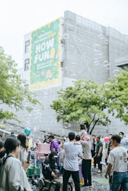 A lively outdoor gathering with many people, including families and children, enjoying themselves in a public space. Soap bubbles float in the air, adding a whimsical touch to the scene. A large colorful banner on a building in the background with the words 'HOW FUN'. People are interacting, some are taking photos, and a child is sitting on an adult's shoulders. The atmosphere is festive and cheerful.