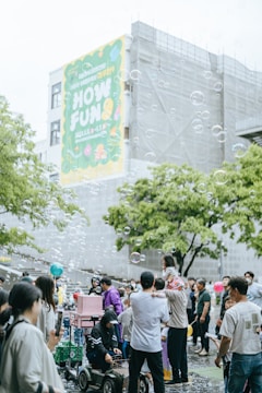A lively outdoor gathering with many people, including families and children, enjoying themselves in a public space. Soap bubbles float in the air, adding a whimsical touch to the scene. A large colorful banner on a building in the background with the words 'HOW FUN'. People are interacting, some are taking photos, and a child is sitting on an adult's shoulders. The atmosphere is festive and cheerful.