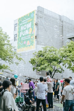 A lively outdoor gathering with many people, including families and children, enjoying themselves in a public space. Soap bubbles float in the air, adding a whimsical touch to the scene. A large colorful banner on a building in the background with the words 'HOW FUN'. People are interacting, some are taking photos, and a child is sitting on an adult's shoulders. The atmosphere is festive and cheerful.