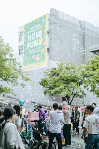 A lively outdoor gathering with many people, including families and children, enjoying themselves in a public space. Soap bubbles float in the air, adding a whimsical touch to the scene. A large colorful banner on a building in the background with the words 'HOW FUN'. People are interacting, some are taking photos, and a child is sitting on an adult's shoulders. The atmosphere is festive and cheerful.
