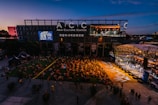 An outdoor event at the Asia Culture Center features a large audience sitting on the grass, watching a live performance on a stage. The stage is brightly lit, contrasting with the darkening evening sky. A large screen displays the performers, and there's signage for the 'World Music Festival' on the buildings.