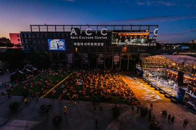 An outdoor event at the Asia Culture Center features a large audience sitting on the grass, watching a live performance on a stage. The stage is brightly lit, contrasting with the darkening evening sky. A large screen displays the performers, and there's signage for the 'World Music Festival' on the buildings.