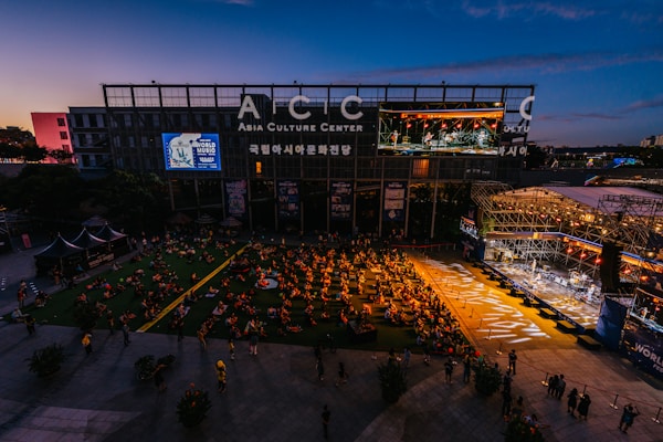An outdoor event at the Asia Culture Center features a large audience sitting on the grass, watching a live performance on a stage. The stage is brightly lit, contrasting with the darkening evening sky. A large screen displays the performers, and there's signage for the 'World Music Festival' on the buildings.