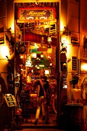 A warmly lit entrance to an Argentine grill, decorated with colorful lights and various objects. The eclectic décor includes hanging lamps, signs, and a red duct. Two people are seen entering the establishment, which is filled with vibrant and inviting lighting.