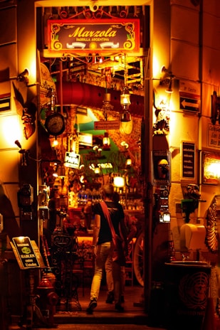 A warmly lit entrance to an Argentine grill, decorated with colorful lights and various objects. The eclectic décor includes hanging lamps, signs, and a red duct. Two people are seen entering the establishment, which is filled with vibrant and inviting lighting.