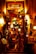 A warmly lit entrance to an Argentine grill, decorated with colorful lights and various objects. The eclectic décor includes hanging lamps, signs, and a red duct. Two people are seen entering the establishment, which is filled with vibrant and inviting lighting.