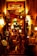 A warmly lit entrance to an Argentine grill, decorated with colorful lights and various objects. The eclectic décor includes hanging lamps, signs, and a red duct. Two people are seen entering the establishment, which is filled with vibrant and inviting lighting.