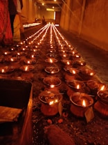 Rows of clay oil lamps are lit and arranged in an orderly fashion on the ground inside a dimly lit corridor. Two people stand on the side, partially visible in traditional attire. The warm glow from the lamps illuminates the space, creating a serene and festive ambiance.