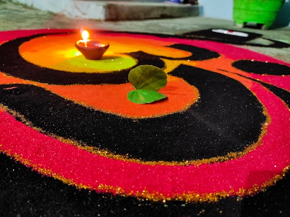 A vibrant design made with colored powders, featuring an earthen lamp placed on it with a lit flame. Two green leaves are situated nearby, adding a natural element to the scene. The design consists of concentric circles in red, orange, and black colors, creating a striking and festive appearance.