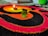 A vibrant design made with colored powders, featuring an earthen lamp placed on it with a lit flame. Two green leaves are situated nearby, adding a natural element to the scene. The design consists of concentric circles in red, orange, and black colors, creating a striking and festive appearance.