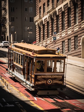 A classic San Francisco cable car is seen traveling along a steep street lined with historic brick buildings. The shadows cast by the buildings create a striking contrast with the sunlit areas.