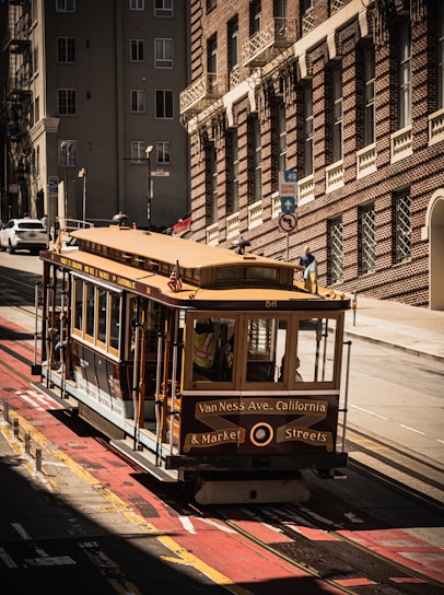 A classic San Francisco cable car is seen traveling along a steep street lined with historic brick buildings. The shadows cast by the buildings create a striking contrast with the sunlit areas.