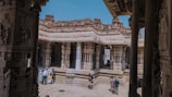 A group of people is exploring a historical temple with intricately carved stone columns and ornate architecture. The temple is partly open to the sky, showcasing a mix of ancient and weathered stone structures. Bright sunlight casts shadows on the stone floor, enhancing the textures of the carvings.