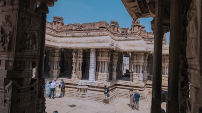 A group of people is exploring a historical temple with intricately carved stone columns and ornate architecture. The temple is partly open to the sky, showcasing a mix of ancient and weathered stone structures. Bright sunlight casts shadows on the stone floor, enhancing the textures of the carvings.