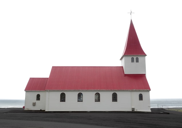A white church with a distinct red roof and steeple stands prominently against an overcast sky. The structure features a series of arched windows along its side and a large cross atop the steeple. The church is situated on a dark, paved surface with the sea visible in the background.