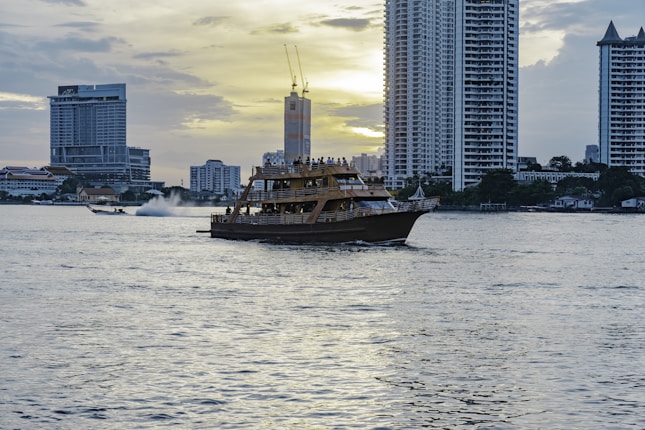 A large wooden boat is cruising on a calm body of water during sunset. Skyscrapers stand tall in the background, their reflections shimmering on the surface. The sky is partially cloudy, with the sun setting behind the buildings, casting a serene glow.