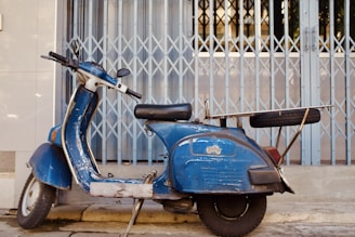 A vintage blue scooter is parked on a concrete surface in front of a metal folding gate. The scooter appears worn, with signs of rust and chipped paint. Mounted on its rear is a metal rack carrying a spare tire.